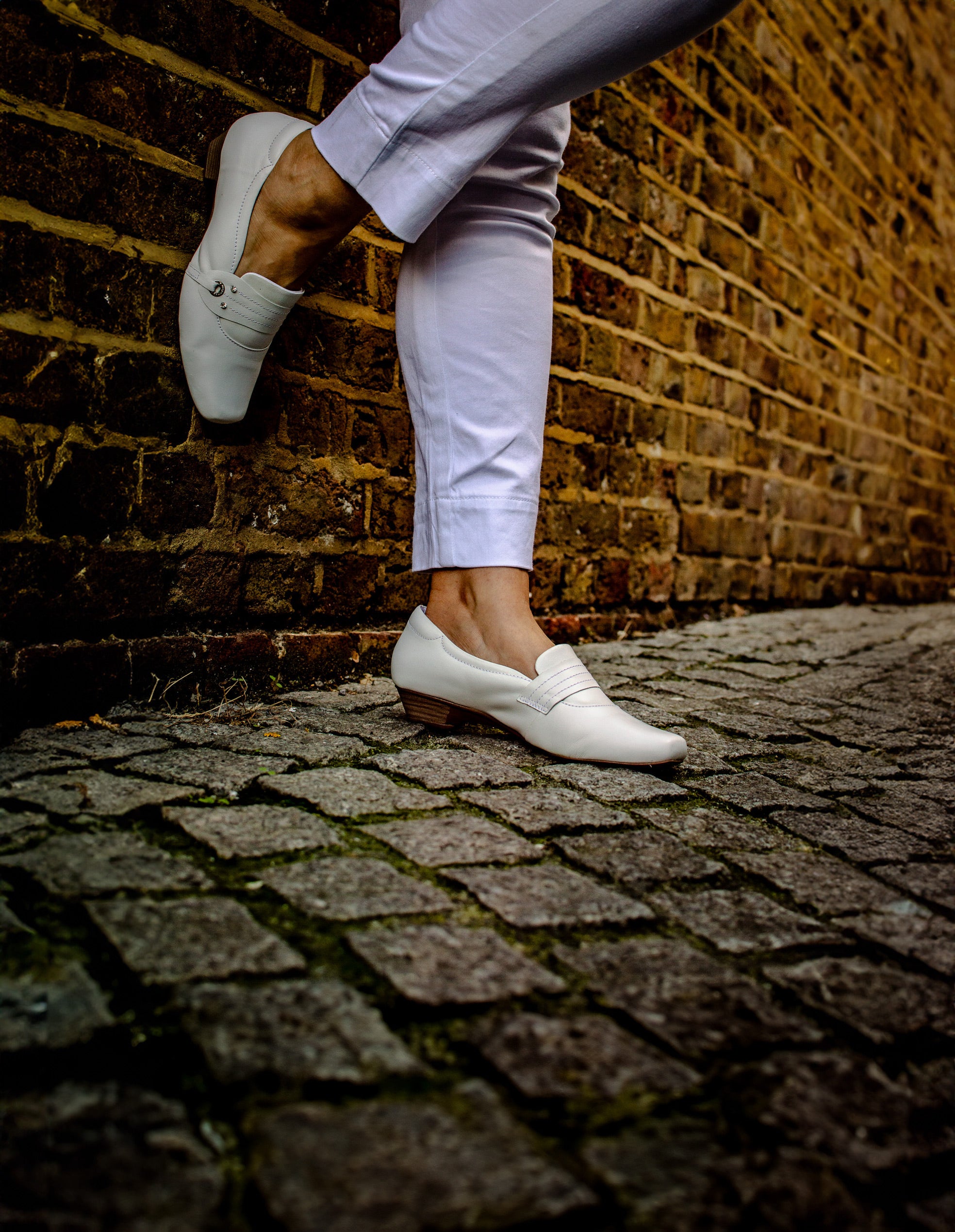 Woman wearing comfortable Terraloom white shoes on cobblestone street with brick wall - urban lifestyle photography